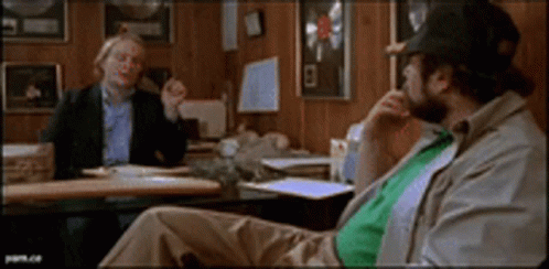 Two Men Are Sitting At A Table Talking To Each Other GIF