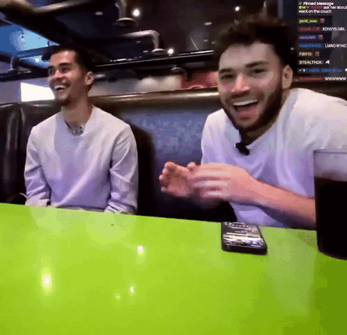 Two Men Are Sitting At A Table With A Green Table Cloth And A Remote Control On It GIF