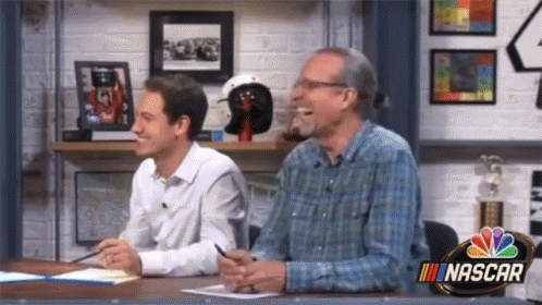 Two Men Are Sitting At A Table With A Nascar Logo On The Wall GIF