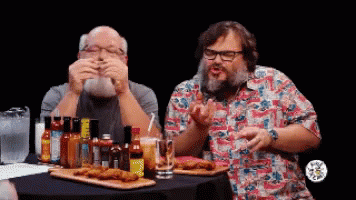 Two Men Are Sitting At A Table With Hot Sauce Bottles GIF