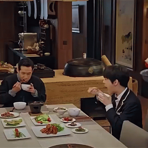 Two Men Are Sitting At A Table With Plates Of Food GIF