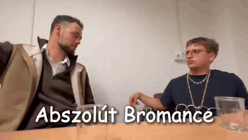 Two Men Are Sitting At A Table With The Words Abszolút Bromance Written On The Bottom GIF