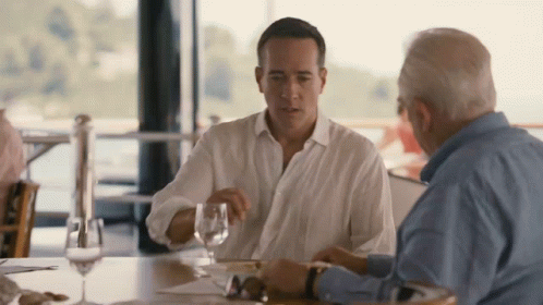 Two Men Are Sitting At A Table With Wine Glasses GIF