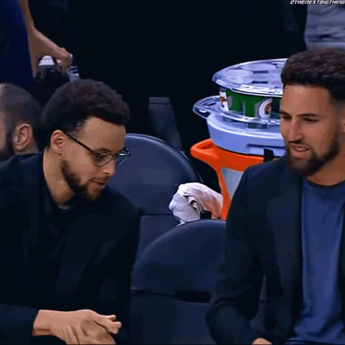 Two Men Are Sitting In A Stadium With A Gatorade Cooler In The Back GIF