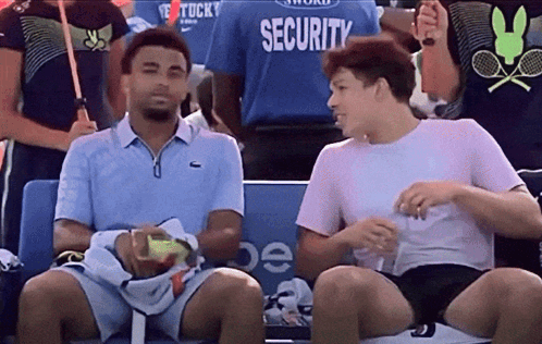 Two Men Are Sitting In A Stadium With A Security Guard In The Background GIF