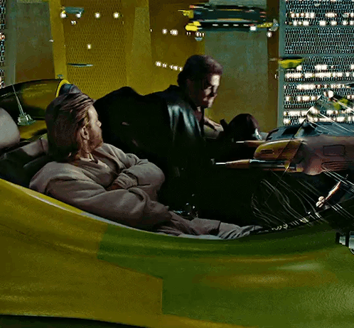 Two Men Are Sitting In A Yellow And Green Vehicle With A City In The Background GIF