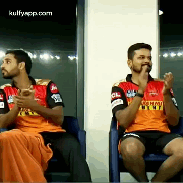 Two Men Are Sitting In Chairs And Clapping Their Hands While Wearing Orange Jerseys GIF