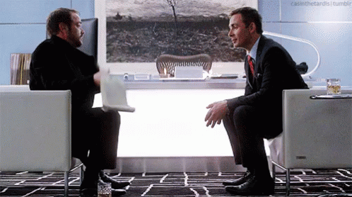 Two Men Are Sitting In Chairs Talking To Each Other GIF