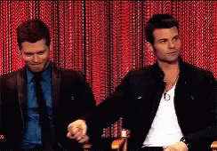 Two Men Are Sitting In Front Of A Red Curtain Holding Hands GIF