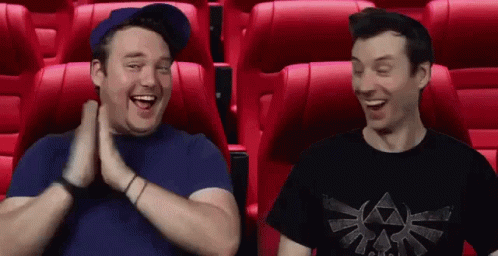 Two Men Are Sitting In Red Seats In A Movie Theater And Laughing . GIF