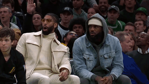 two men are sitting in the stands at a basketball game and one has the letter l on his shirt