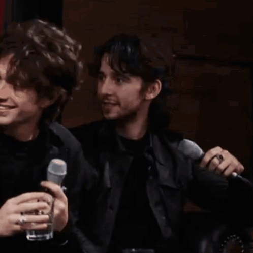 Two Men Are Sitting Next To Each Other Holding Microphones And Drinking . GIF