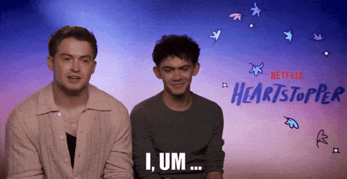 Two Men Are Sitting Next To Each Other In Front Of A Netflix Advertisement For Heartstopper GIF