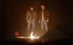 Two Men Are Standing Around A Fire In The Dark . GIF