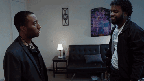 Two Men Are Standing In A Living Room With A Purple Poster On The Wall GIF