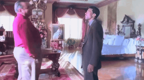 Two Men Are Standing In A Room With A Couch And A Painting On The Wall GIF