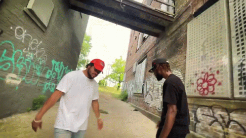 Two Men Are Standing In Front Of A Brick Wall With Graffiti On It Including One That Says Check GIF