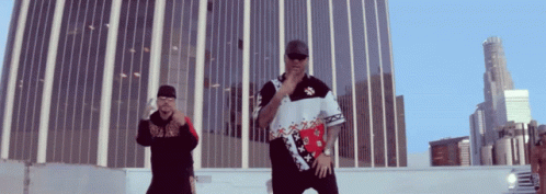 Two Men Are Standing In Front Of A Tall Building And One Has A Shirt That Says ' Tommy Hilfiger ' On It GIF