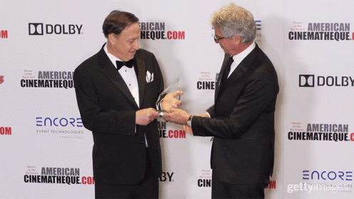 Two Men Are Standing In Front Of A Wall That Says Dolby On It GIF