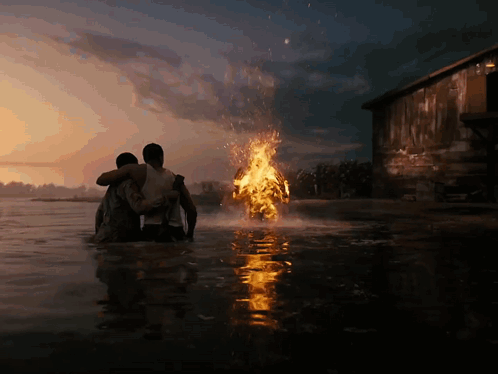 Two Men Are Standing In The Water Looking At A Fire Coming Out Of The Water GIF