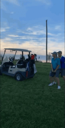 Two Men Are Standing Next To A Golf Cart On A Grassy Field GIF