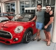 Two Men Are Standing Next To A Red Car In Front Of A Car Dealership . GIF