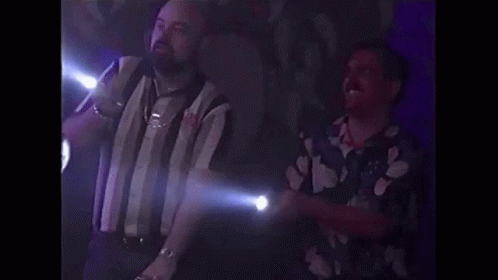 Two Men Are Standing Next To Each Other In A Dark Room . One Of The Men Is Holding A Flashlight . GIF
