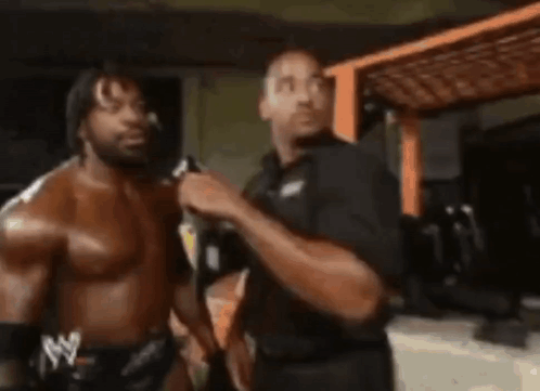 Two Men Are Standing Next To Each Other In A Room . One Of The Men Is Wearing A Shirtless Wrestling Outfit . GIF