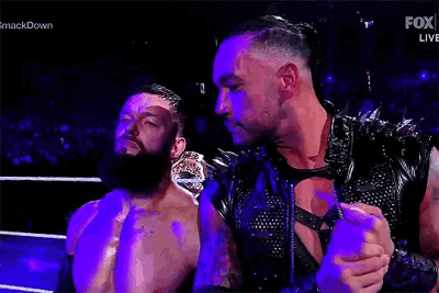 Two Men Are Standing Next To Each Other In A Wrestling Ring . One Of The Men Is Wearing A Leather Vest . GIF