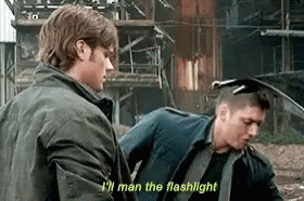 Two Men Are Standing Next To Each Other In Front Of A Building . One Of The Men Is Holding A Flashlight . GIF