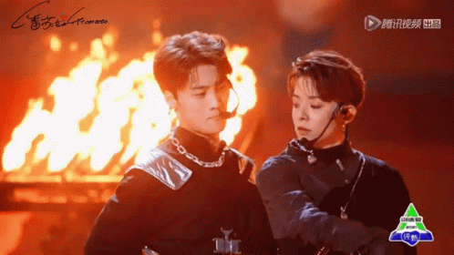 Two Men Are Standing Next To Each Other In Front Of A Fire With A Green Logo On The Bottom Right Corner GIF