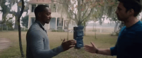 Two Men Are Standing Next To Each Other In Front Of A House . One Of The Men Is Holding A Stack Of Boxes . GIF