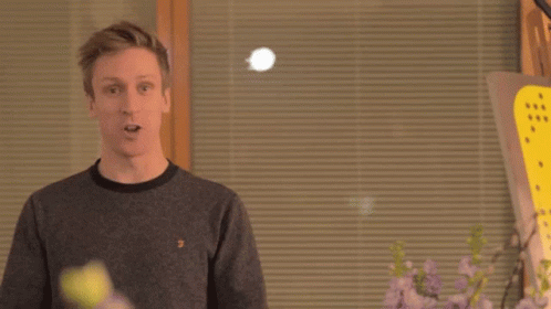Two Men Are Standing Next To Each Other In Front Of A Window With Blinds And Flowers . GIF