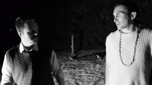 Two Men Are Standing Next To Each Other In The Dark And One Has A Necklace Around His Neck GIF