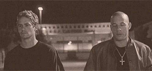 Two Men Are Standing Next To Each Other On A Street At Night . One Of The Men Is Wearing A Cross Necklace . GIF