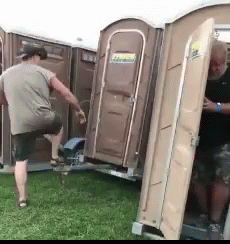 Two Men Are Standing Next To Portable Toilets In A Grassy Field . GIF