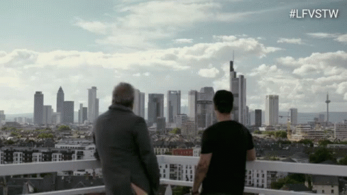 Two Men Are Standing On A Balcony Looking At A City Skyline With The Hashtag #Lfvstw GIF