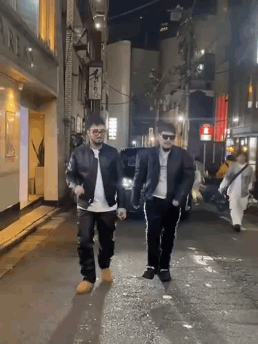 Two Men Are Walking Down A City Street At Night GIF