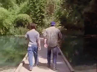 Two Men Are Walking On A Bridge Over A River . GIF