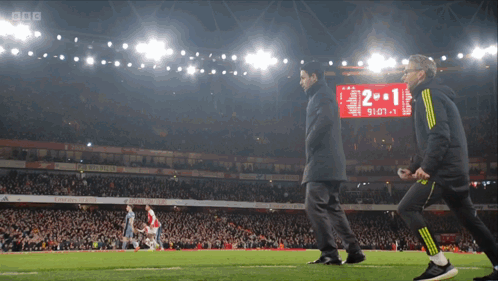 Two Men Are Walking On A Soccer Field With A Scoreboard That Says 2 1 GIF