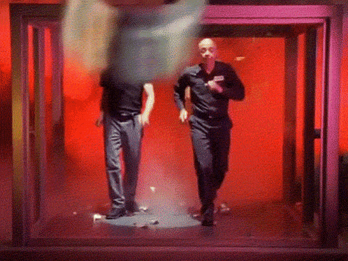 Two Men Are Walking Through A Doorway In A Dark Room GIF
