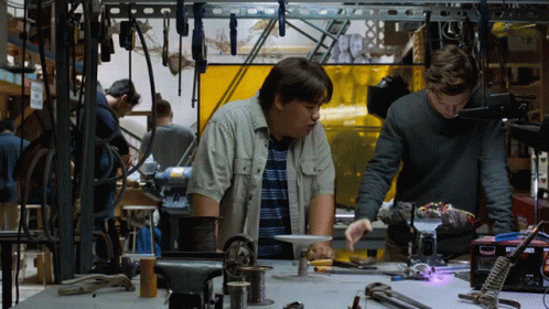 Two Men Are Working On A Project In A Workshop And One Of Them Is Looking At A Box That Says Welding GIF