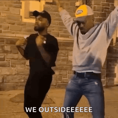 Two Men Dancing Arms Raised Brick Building GIF