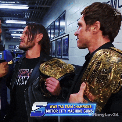 Two Men Holding Wrestling Belts In Front Of A Wwe Tag Team Champions Motor City Machine Guns Banner GIF