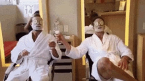 Two Men In Bathrobes With Face Masks On Their Faces Are Sitting In A Chair . GIF