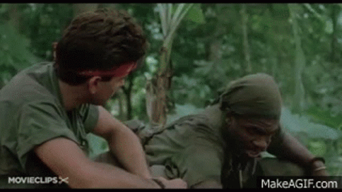 Two Men In Military Uniforms Are Sitting Next To Each Other In The Jungle Smoking Cigarettes . GIF