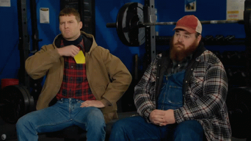 Two Men In Plaid Shirts And Overalls Are Sitting In A Gym One Is Holding A Yellow Card GIF
