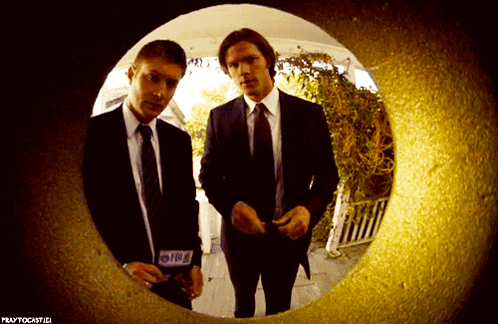 Two Men In Suits And Ties Are Looking Through A Hole In A Wall That Says Praytocastici GIF