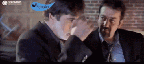 Two Men In Suits And Ties Are Talking In Front Of A Brick Wall With A Blue Sign That Says Pichuka GIF