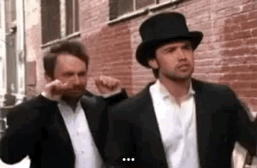 Two Men In Suits And Top Hats Are Standing Next To Each Other On A Sidewalk . GIF